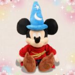Une peluche Mickey déguisé en magicien avec un grand chapeau bleu, une robe rouge et des chaussures marron, posée sur un fond clair décoré d’étoiles et de cœurs.