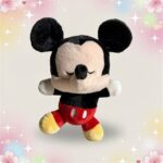 Une peluche Mickey aux yeux fermés, assise avec les bras ouverts, portant un short rouge et des chaussures jaunes sur un fond pastel décoré d’étoiles et de cœurs.