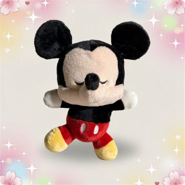 Une peluche Mickey aux yeux fermés, assise avec les bras ouverts, portant un short rouge et des chaussures jaunes sur un fond pastel décoré d’étoiles et de cœurs.