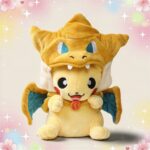 Une peluche Pikachu déguisé en Dracaufeu, avec un bonnet orange à tête de dragon, des petites ailes bleues et un fond doux décoré de cœurs et d’étoiles.