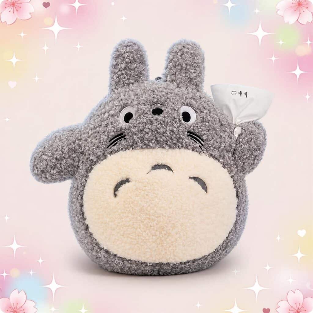 Une peluche Totoro grand format et son baluchon, toute douce et grise, avec un grand ventre clair, posée sur un fond pastel avec des petites fleurs.