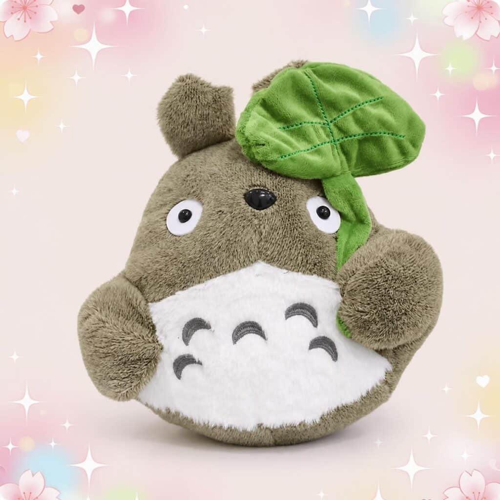 Une peluche Totoro tenant une grande feuille verte, avec un ventre blanc et un grand regard rond, sur un fond pastel décoré de petites étoiles et fleurs.