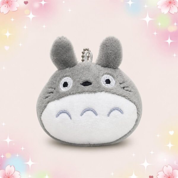 Une peluche Totoro version miniature porte-clés grise et douce, avec une petite chaîne en métal, posée sur un fond rose clair décoré d’étoiles et de fleurs.