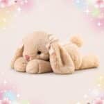 Une peluche lapin Kawaii timide transformable en oreiller douillet beige, toute molle avec de longues oreilles, un petit nez rose et des nœuds à carreaux, posée sur un fond pastel avec des fleurs et des étoiles.