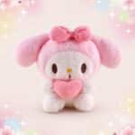 Une peluche Sanrio My Melody rose et blanche, avec un gros nœud sur la tête et un cœur rose dans les mains, sur un fond pastel décoré d’étoiles et de petits cœurs.