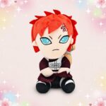 Une peluche Gaara de l'anime Naruto position assise, avec les cheveux rouges, de grands yeux bleus et un petit costume, posée sur un fond rose décoré d’étoiles et de fleurs.