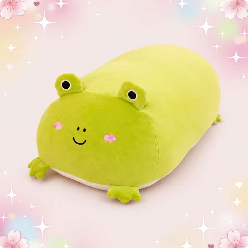 Une peluche grenouille squishy toute en longueur verte, toute douce et rigolote, posée sur un fond rose avec des petites fleurs et des étoiles brillantes.