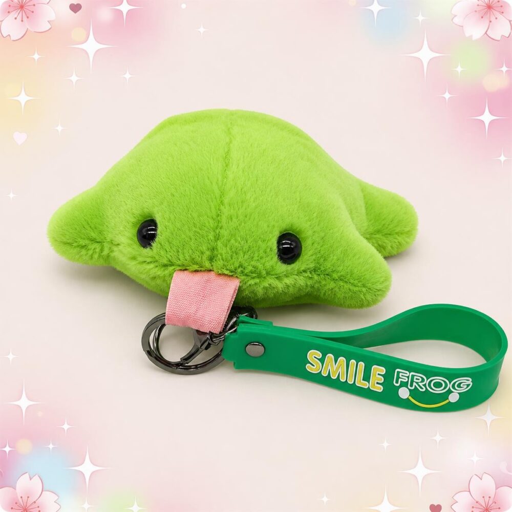 Une peluche grenouille tirant la langue format porte-clés verte, avec des yeux noirs brillants et une lanière verte, posée sur un fond rose pastel avec des cœurs et des étoiles.