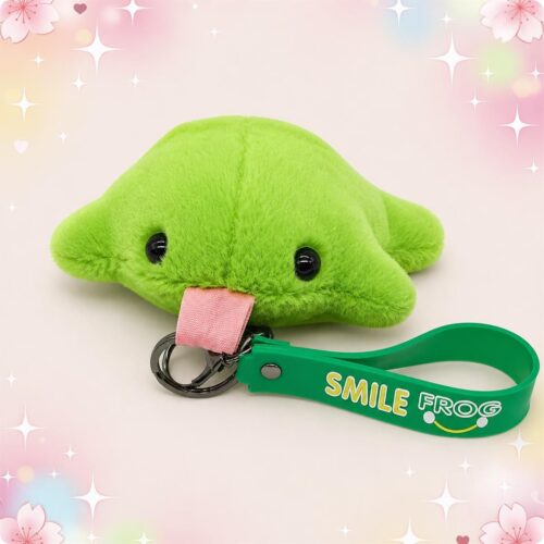 Une peluche grenouille tirant la langue format porte-clés verte, avec des yeux noirs brillants et une lanière verte, posée sur un fond rose pastel avec des cœurs et des étoiles.