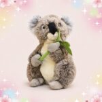 Une peluche Koala tenant une feuille eucalyptus dans les pattes, assise sur un fond rose doux avec des étoiles et des fleurs.