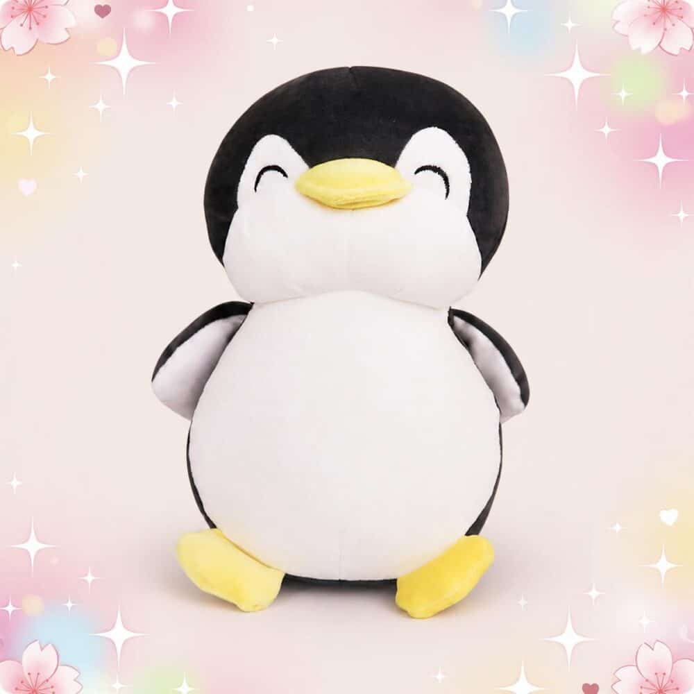 Une peluche pingouin grand format et squishy toute douce, en noir et blanc avec un bec et des pattes jaunes, posée sur un fond rose pastel décoré d’étoiles et de cœurs.