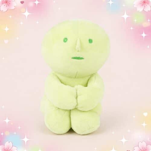 Une peluche Smiski assis verte pastel, avec un petit visage simple et les bras croisés, posée sur un fond rose doux avec des étoiles brillantes.
