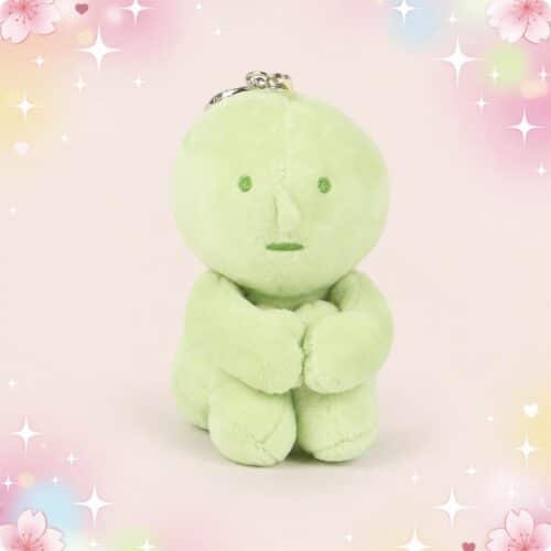 Une peluche Smiski assis format porte-clés verte, toute ronde, avec un petit visage, posée sur un fond rose pastel décoré d’étoiles et de fleurs.