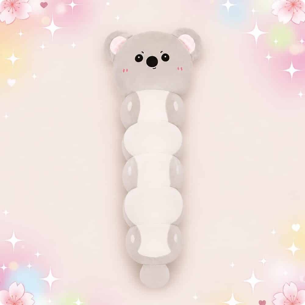 Une peluche Koala grise en forme de long coussin doux, avec un visage mignon et des parties blanches, posée sur un fond rose pastel décoré d’étoiles et de petits cœurs.