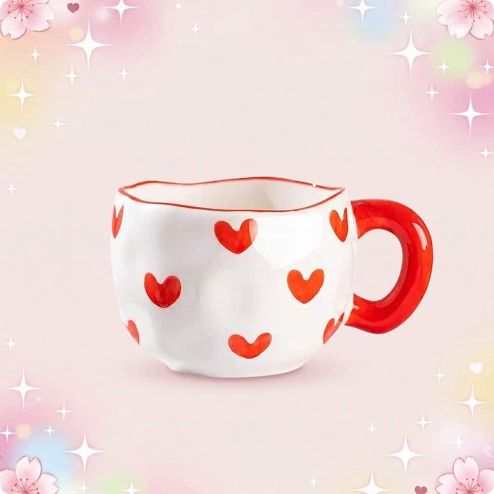 Une tasse kawaii à motif coeurs blanche avec des coeurs rouges et une anse rouge, posée sur un fond rose doux et scintillant.