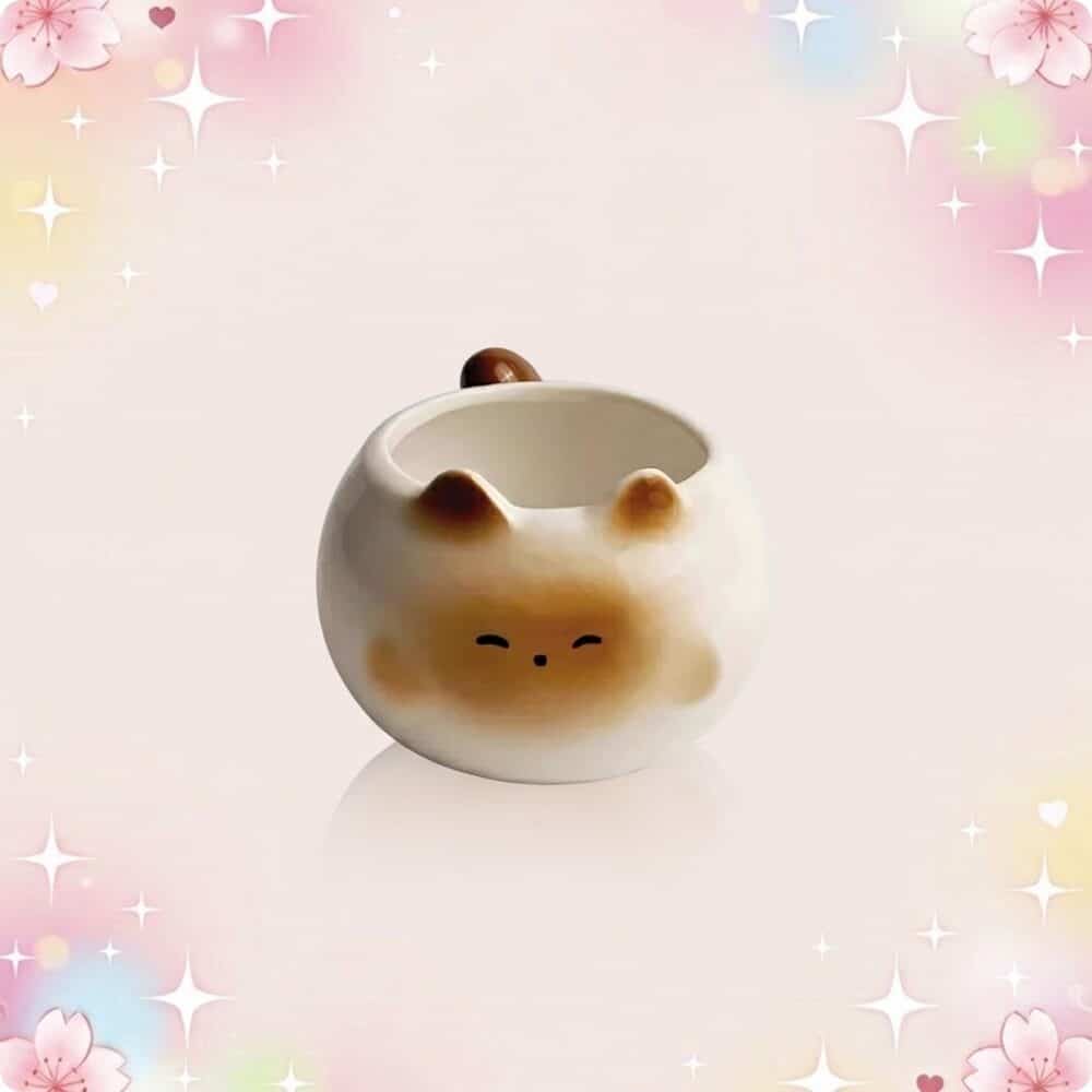 Une tasse kawaii chat mignon ronde en céramique, avec une petite tête de chat et des oreilles, posée sur un fond rose pastel avec des étoiles scintillantes.