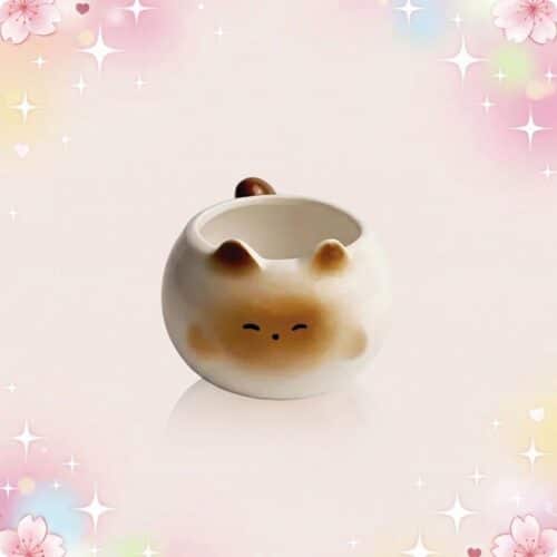 Une tasse kawaii chat mignon ronde en céramique, avec une petite tête de chat et des oreilles, posée sur un fond rose pastel avec des étoiles scintillantes.