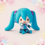 Une figurine kawaii Hatsune Miku assise, avec de longs cheveux bleus en queues de cheval, sur un fond pastel décoré d’étoiles et de petits cœurs.