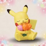 Une figurine kawaii Pikachu tenant des fleurs, assise avec les yeux fermés et un fond pastel décoré de cœurs et d’étoiles.