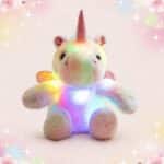 Une peluche licorne lumineuse en tissu doux, assise et brillant de couleurs arc-en-ciel, avec une corne rose et un fond rose décoré d’étoiles et de petits cœurs.