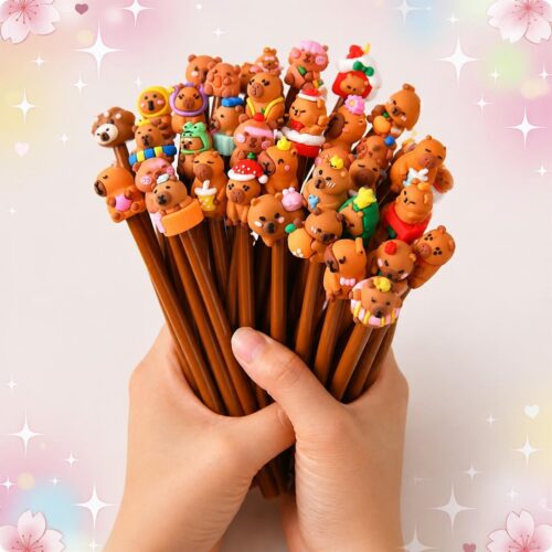 Des mains tiennent un lot de vingt cinq stylo Kawaii capybara noir, avec de petites figurines de capybaras sur le dessus, sur un fond rose pastel décoré d’étoiles et de cœurs.