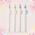 Quatre stylo Kawaii chaton aimanté à pression rétractable blancs, avec un petit chaton coloré au sommet et sur le côté, posés sur un fond rose pastel décoré de cœurs et d’étoiles.