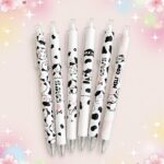 Plusieurs stylo Kawaii vache à pointe extra fine posés sur un fond rose décoré d’étoiles, avec des motifs de vaches noires et blanches mignonnes.