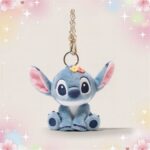 Un porte-clé peluche Stitch bleu, avec de grandes oreilles roses et une petite chaîne métallique, posé sur un fond pastel décoré d’étoiles et de fleurs.