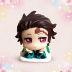 Une petite Figurine Gachapon Tanjiro de Demon Slayer assise, avec de grands yeux roses et un socle blanc, sur un fond pastel avec des fleurs et des étoiles brillantes.