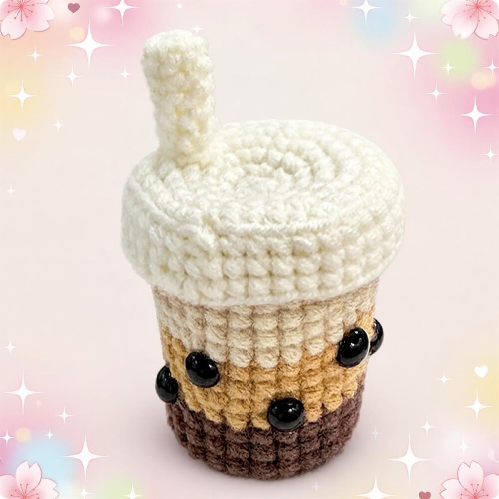 Un kit amigurumi bubble tea représentant une petite boisson en crochet avec paille, crème blanche et perles noires, sur un fond rose décoré d’étoiles et de fleurs.