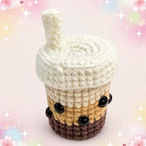 Un kit amigurumi bubble tea représentant une petite boisson en crochet avec paille, crème blanche et perles noires, sur un fond rose décoré d’étoiles et de fleurs.
