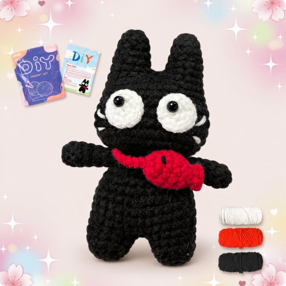 Un kit amigurumi chat noir avec un poisson rouge montrant une petite peluche noire aux grands yeux blancs, avec le fil et les accessoires de crochet autour.