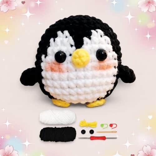 Un kit amigurumi pingouin avec un petit pingouin crocheté, des pelotes de laine blanche et noire, des yeux, un crochet et des accessoires sur fond pastel décoré.