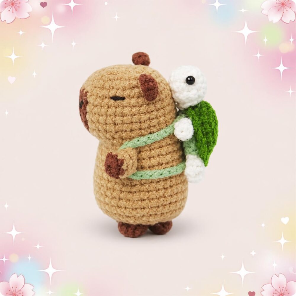 Un petit capibara en crochet portant une tortue sur son dos, avec une carapace verte et un fond pastel décoré d’étoiles et de fleurs.