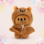 Une mini peluche SKZOO Han Quokka brune en forme de petit animal, avec une capuche à oreilles et un petit sourire, sur fond pastel décoré d’étoiles et de fleurs.