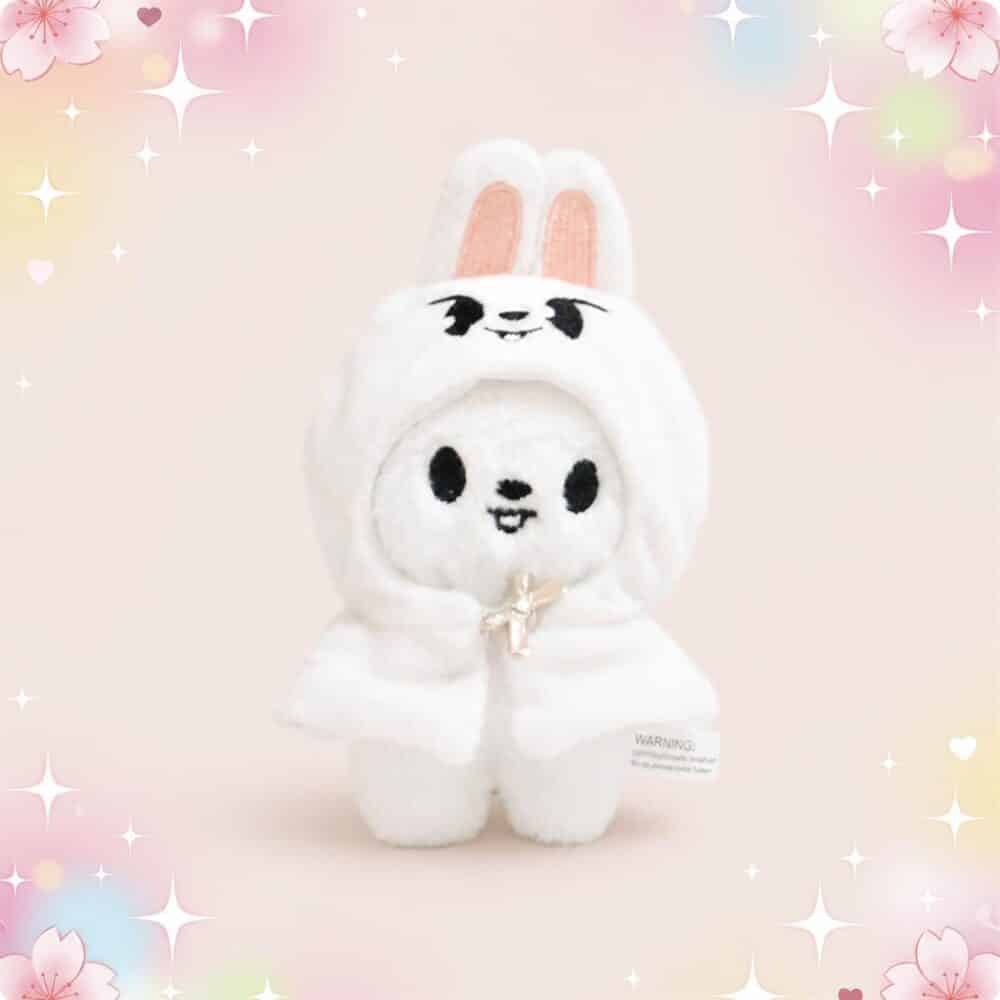 Une mini peluche SKZOO Leebit blanche en costume de lapin, avec de grandes oreilles et un petit sourire, sur un fond rose doux et scintillant.
