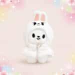Une mini peluche SKZOO Leebit blanche en costume de lapin, avec de grandes oreilles et un petit sourire, sur un fond rose doux et scintillant.