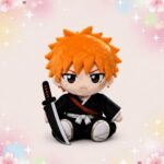 Une peluche anime Bleach Ichigo assise, avec cheveux orange, tenue noire de samouraï et petit sabre, sur fond pastel décoré d’étoiles et de fleurs.