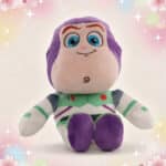 Une peluche Disney Buzz l'Eclair de Toy Story assise, avec combinaison blanche, verte et violette, sur un fond clair décoré de petites étoiles et de cœurs.