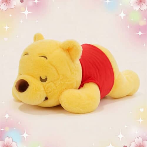 Une peluche Disney Winnie l'Ourson endormi jaune et rouge, couchée sur le ventre avec les yeux fermés sur un fond doux et pastel.