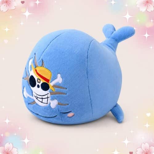 Une peluche manga Laboon One Piece bleue en forme de baleine, avec le drapeau de pirate sur le visage, posée sur un fond pastel décoré d’étoiles et de fleurs.
