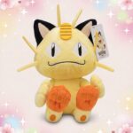 Une peluche Pokemon Miaouss beige assise, avec de grandes oreilles noires, des yeux en amande et de petites pattes orange, sur un fond rose décoré d’étoiles et de cœurs.