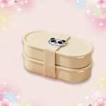 Un bento kawaii à motif panda beige à deux compartiments, fermé par une sangle avec un petit panda, posé sur un fond rose doux et scintillant.