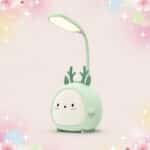 Une Lampe Kawaii pliable avec son petit élan vert lumineux, en forme de petit personnage vert avec une tête flexible allumée, sur fond rose avec des étoiles scintillantes.