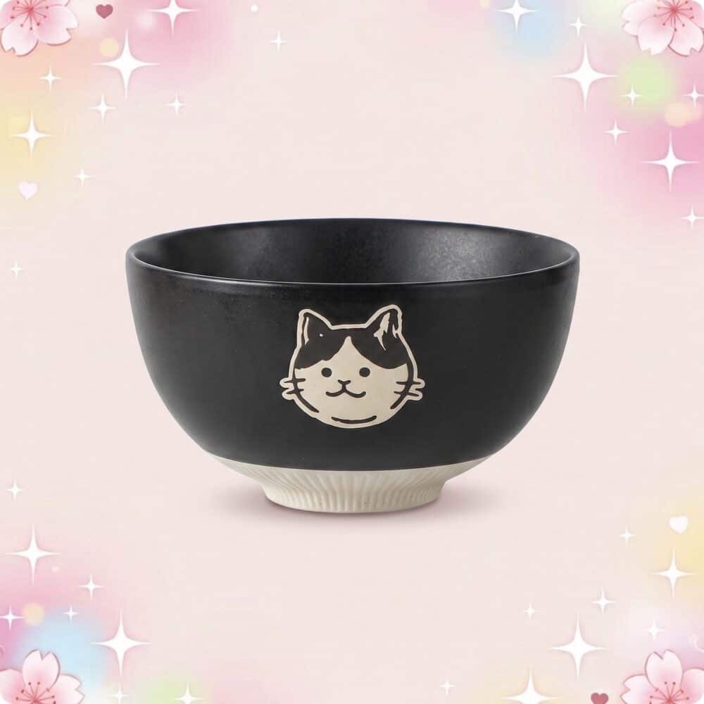 Un bol noir de vaisselle kawaii bol motif animaux mignons, avec un visage de chat blanc et noir, posé sur un fond rose pastel avec des étoiles et des fleurs.