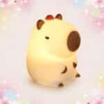 Une Veilleuse Kawaii en capybara à trois modes de luminosité, en forme de petit capybara lumineux, sur un fond rose pastel avec des étoiles et des fleurs.