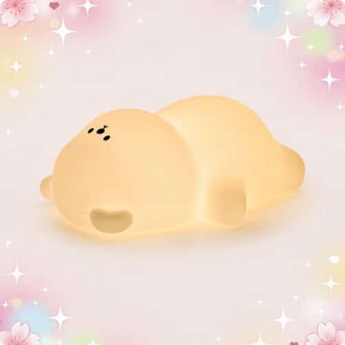 Une douce Veilleuse Kawaii en ourson allongé sur le ventre, en forme de petit ours jaune qui brille, sur un fond pastel avec des étoiles et des cœurs.