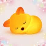 Une Veilleuse Kawaii en renard à extinction automatique en forme de petit renard endormi, qui brille d’une lumière jaune douce sur fond rose décoré d’étoiles et de fleurs.