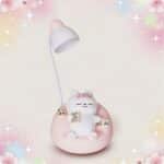 Une lampe kawaii en forme de chat pour bureau étudiant blanche et rose, avec un petit chat assis sur sa base, dans un décor doux et lumineux.