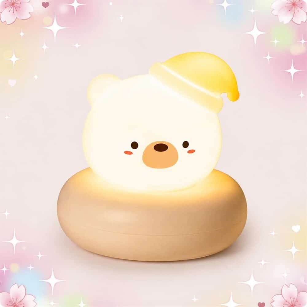 Une Veilleuse Kawaii en ours diffusant une lueur douce en forme d’ours blanc avec un petit bonnet jaune, posée sur une base ronde beige, sur fond rose pastel avec étoiles et cœurs.